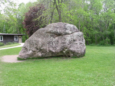 [A huge rock sits in the middle of a grassy patch. On the left edge of the rock is a dirt section where the grass has been worn away. There is a building off to the left with a path leading to it.]